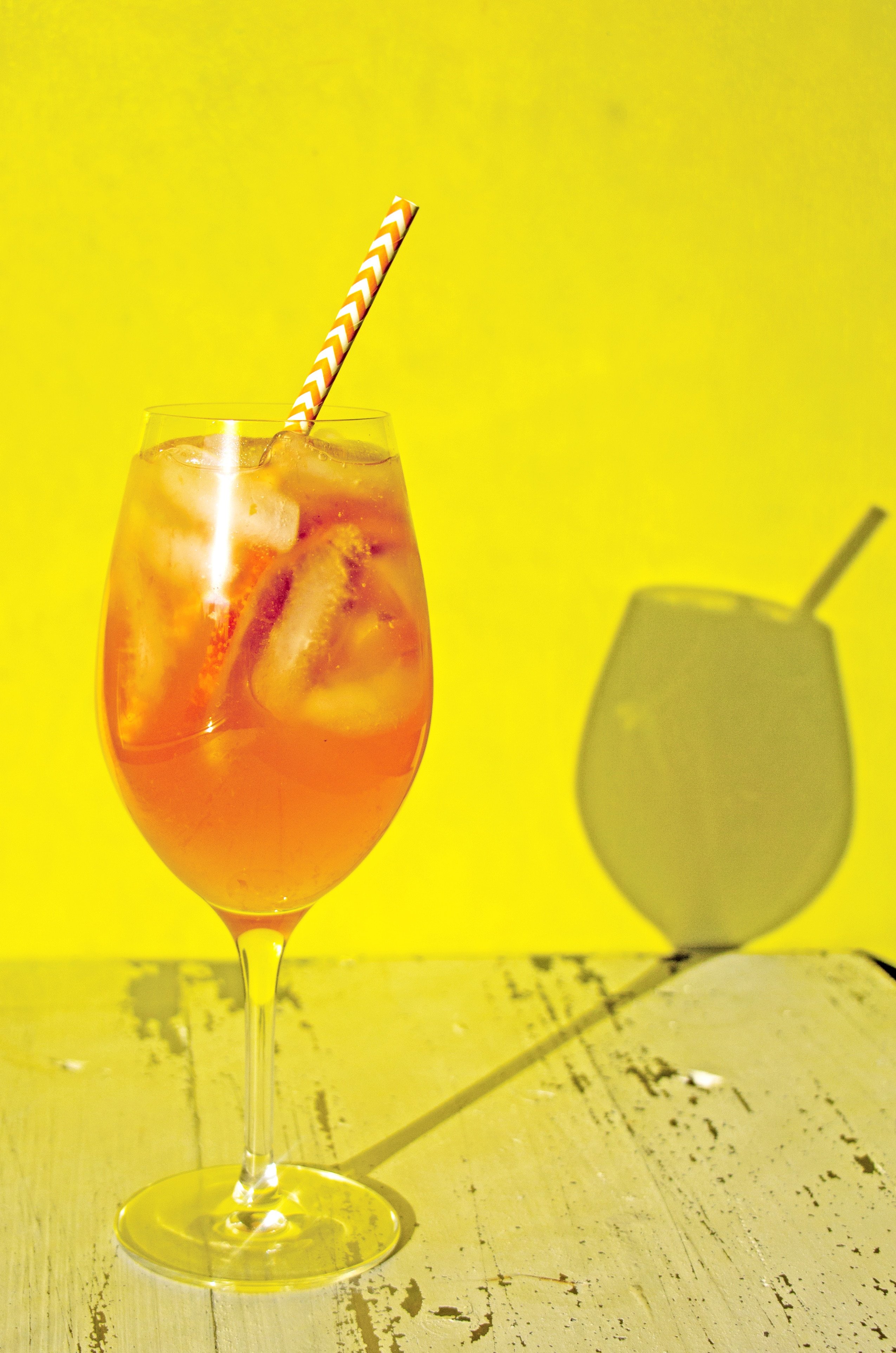 Long-stemmed glass of Orange White Wine Campari Sangria casting a shadow onto a yellow wall.