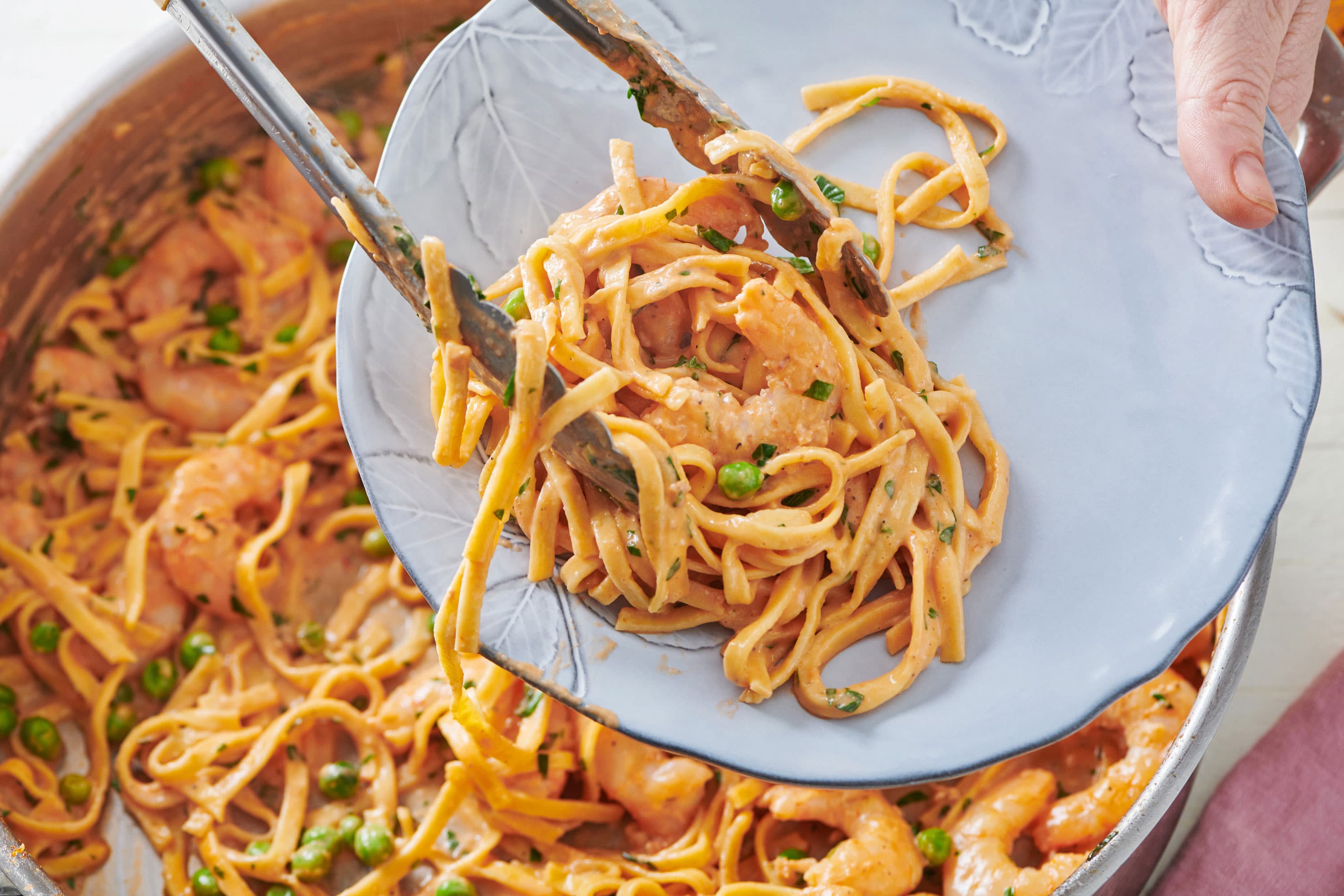 Tongs serving fresh linguine with shrimp and peas in a pink cream sauce onto a plate.