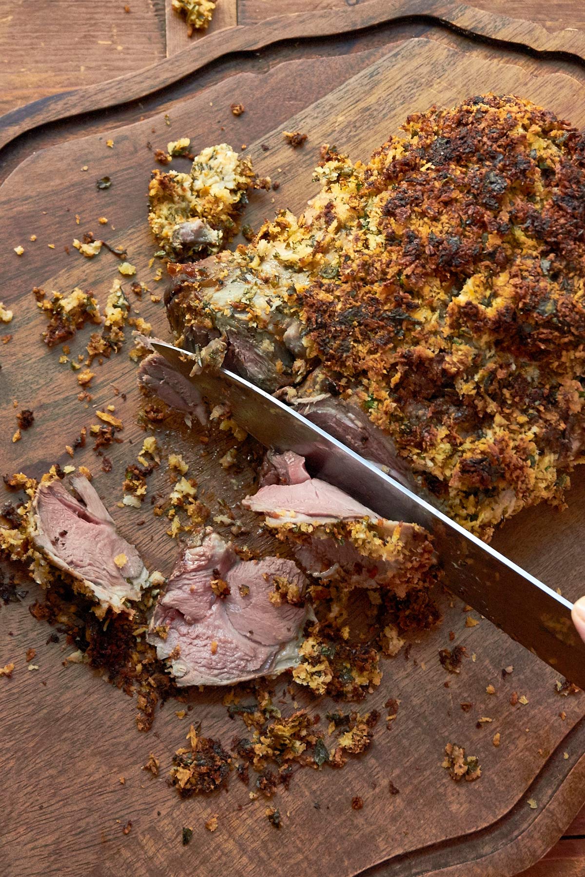 Knife slicing an Herbed Boneless Leg of Lamb with Mustard Crust on a wooden surface.