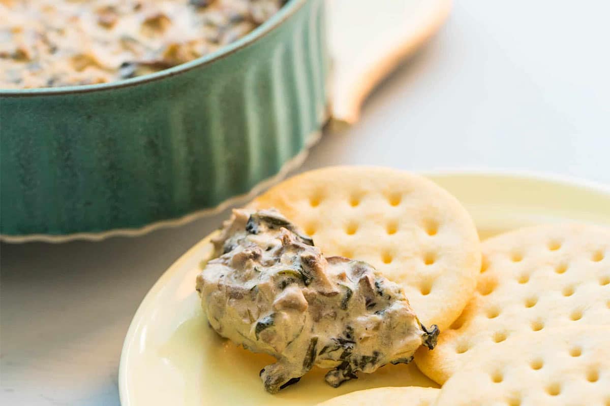 Hot spinach and goat cheese hot dip on cracker in front of green dish.