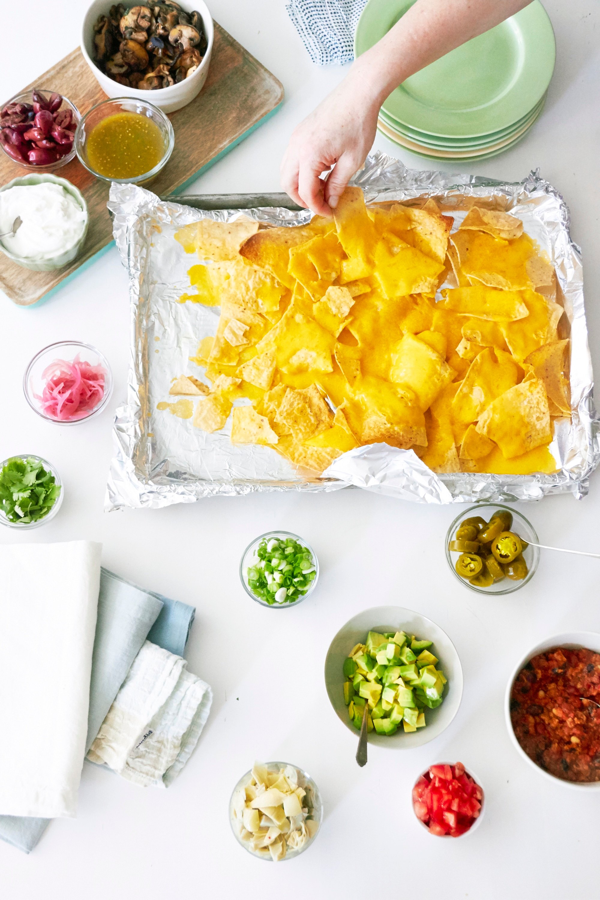 Woman taking a nacho from a baking sheet with nachos on it, surrounded by various topping choices.