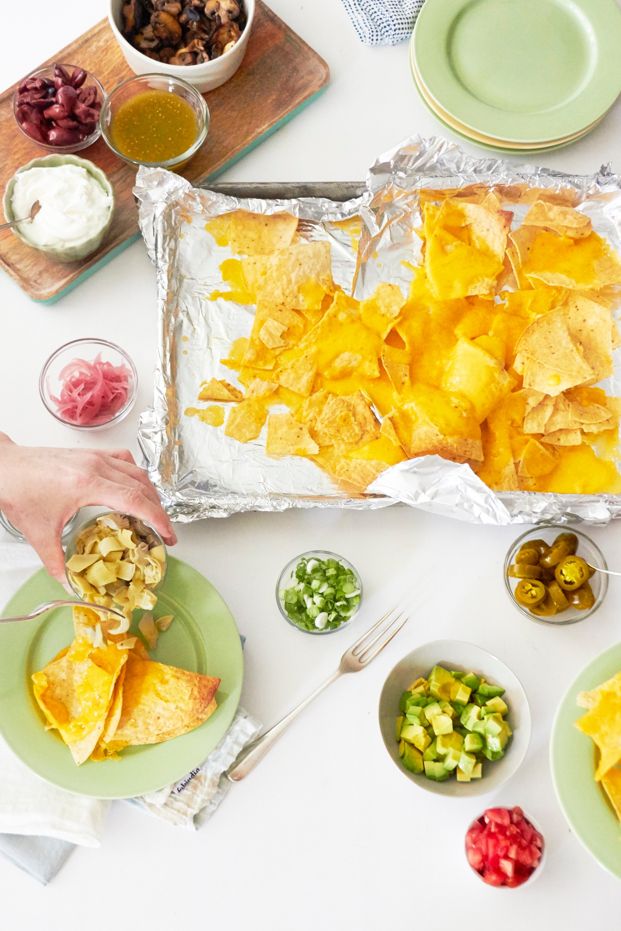 Woman adding some diced avocado hearts to a plate of nachos, with more nachos and toppings nearby