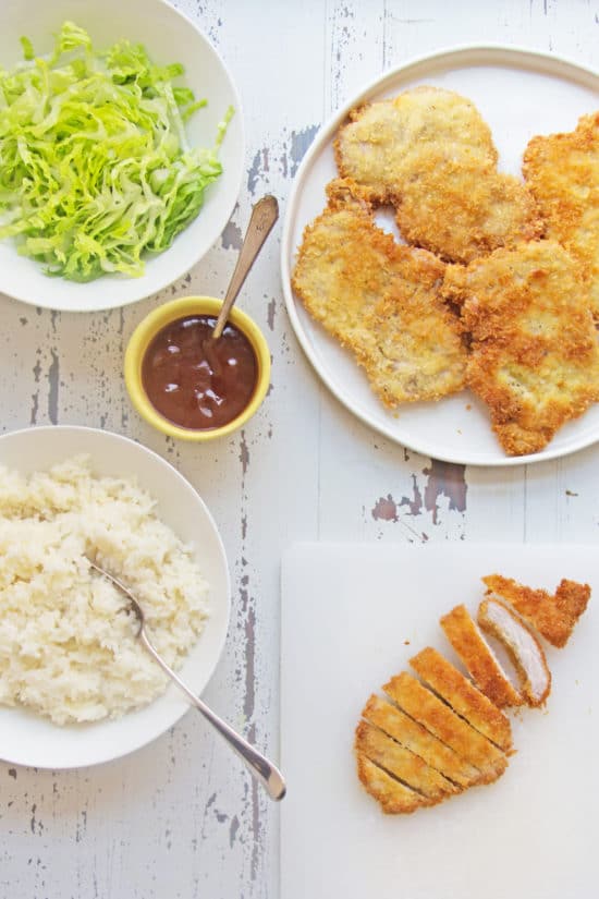 Breaded chicken cutlets, rice, and katsu sauce on a white, wooden table.