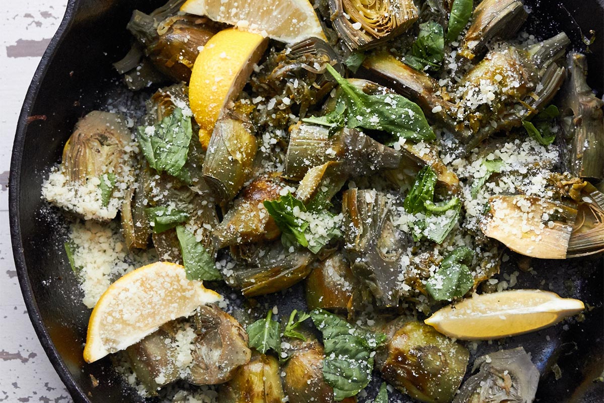 Braised baby artichokes, leeks, and capers with lemon wedges in skillet.