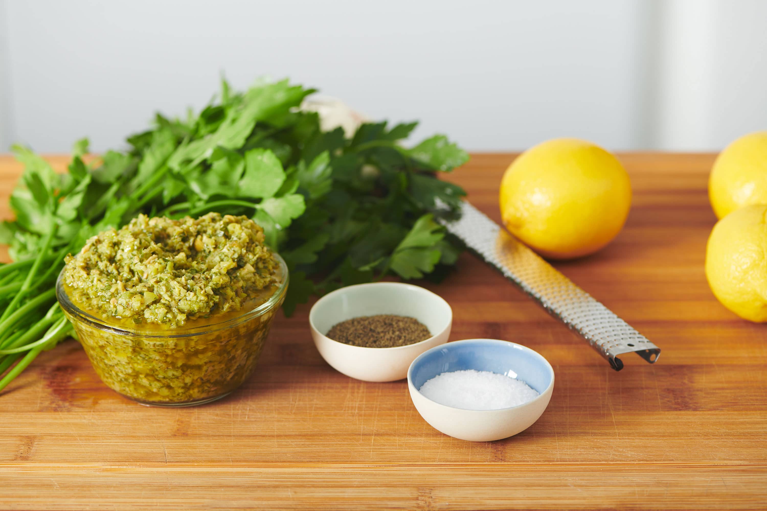 Green Olive Tapenade in a bowl on a wooden counter with ingredients surrounding it.