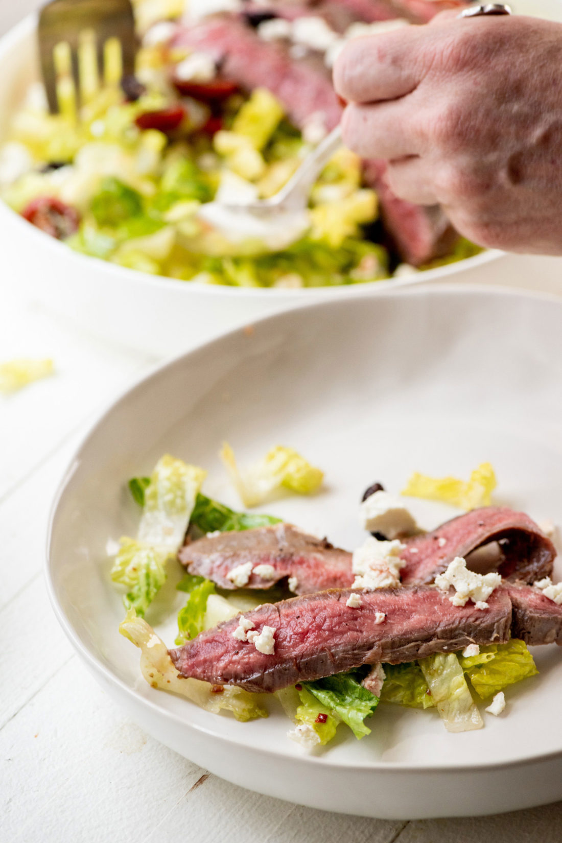 Person serving Greek Salad with Flank Steak onto a plate.