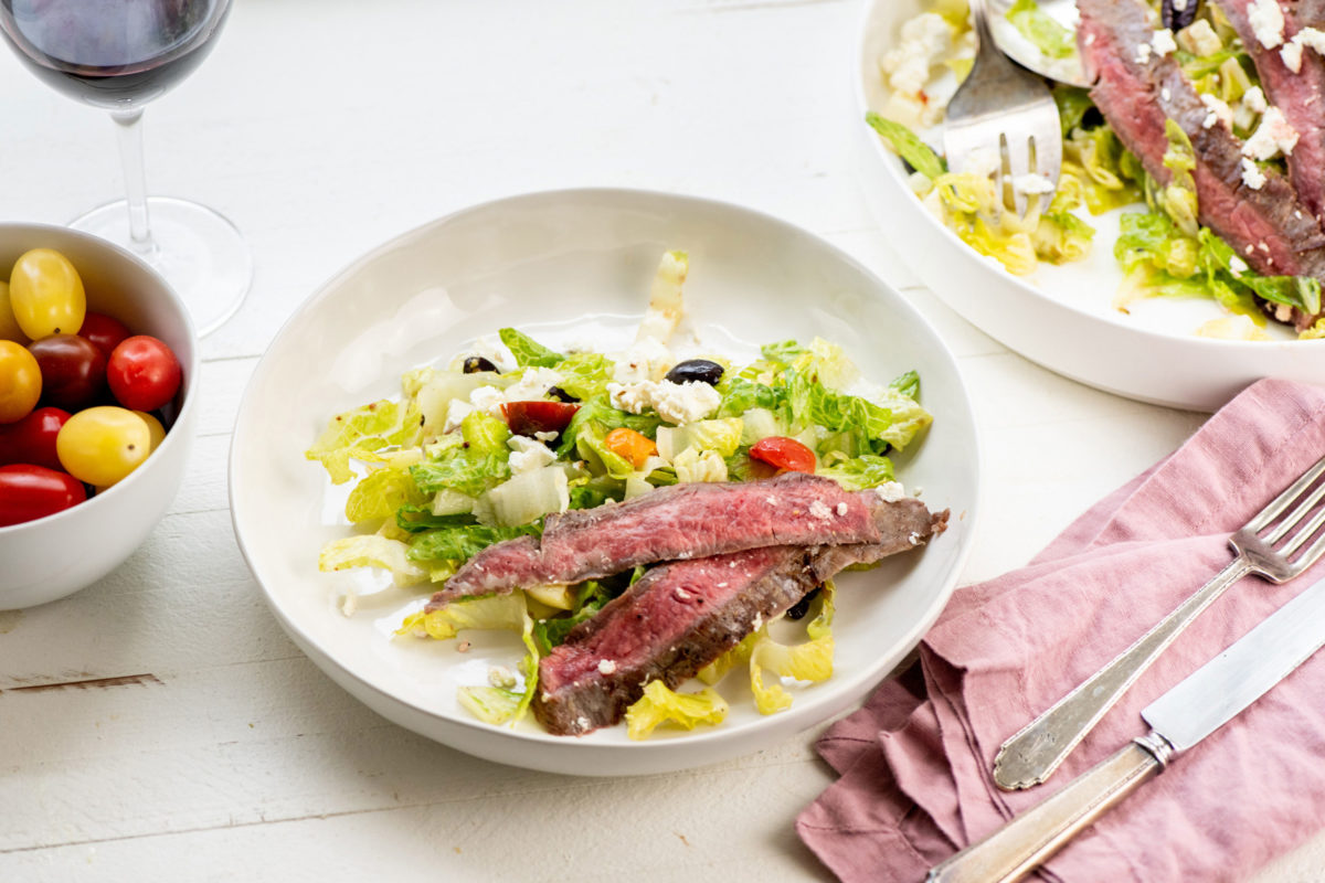Plate of Greek Salad with Flank Steak on a white, wooden table.