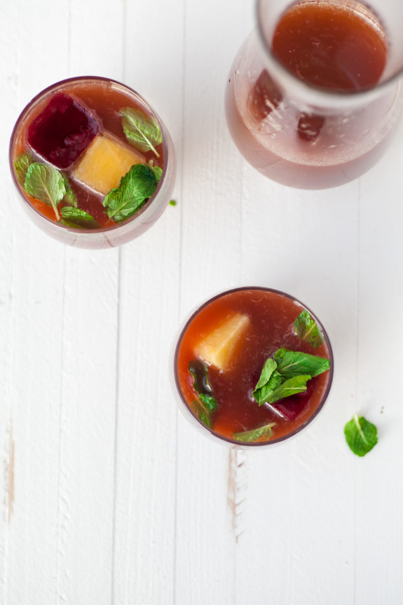 Two glasses of Pomegranate Orange Mocktail with Mint set on a white, wooden table.