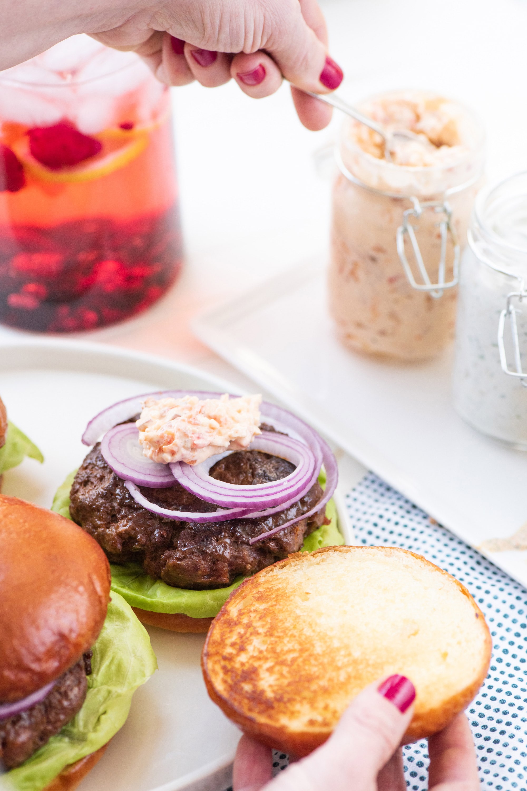Woman scooping pimento cheese spread from glass jar onto burger.