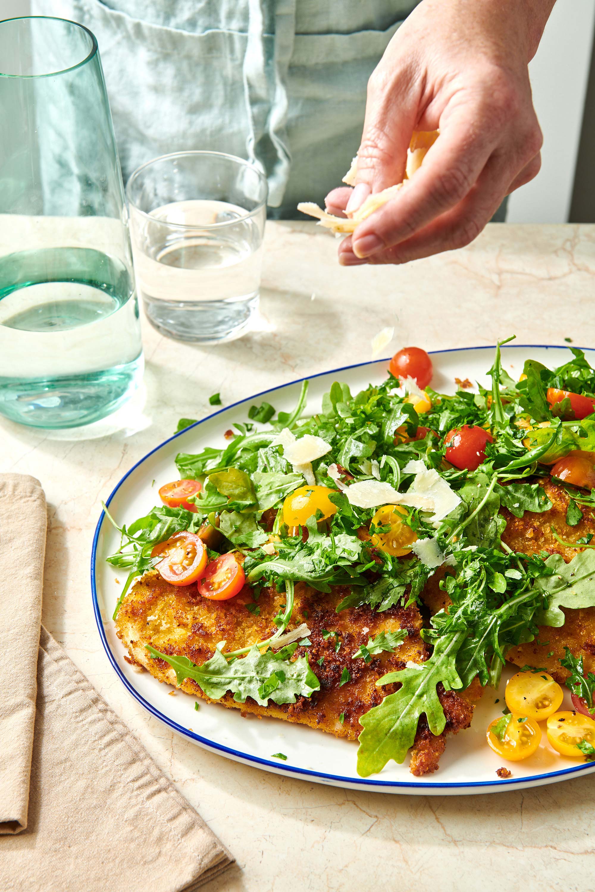 Woman sprinkling shaved parmesan onto a platter of Chicken Milanese topped with salad.