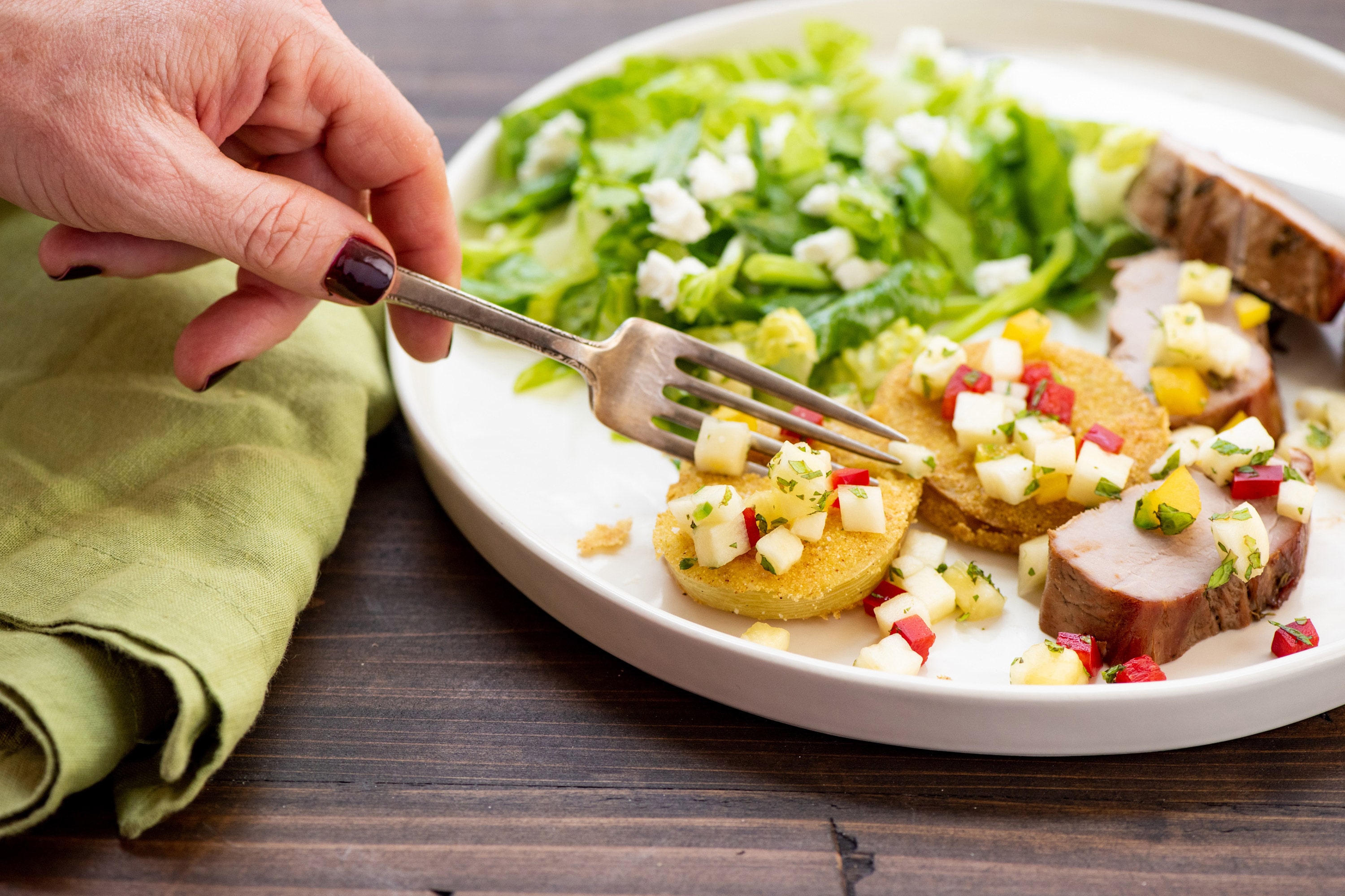 Woman slicing fried green tomato with fork on plate with pork tenderloin and salad.