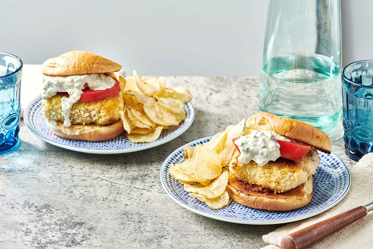 Two fried fish sandwiches with tartar and tomatoes and chips on blue plates.