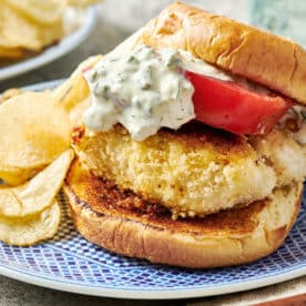 Homemade fried fish sandwich topped with tartar and tomato on blue plate with chips.