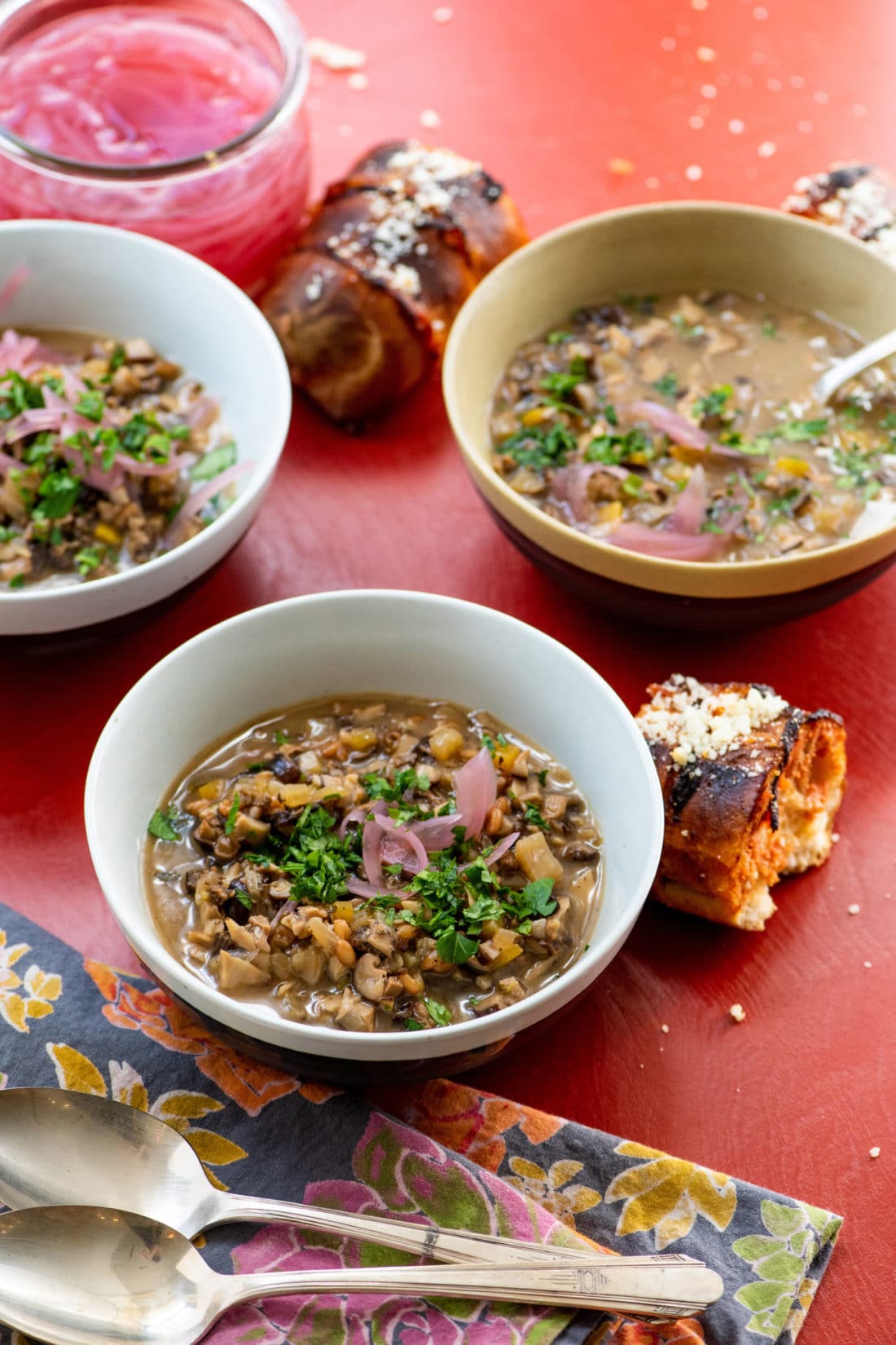 Bowls of Mushroom Spelt Soup topped with pickled onions and parsley on a red table.