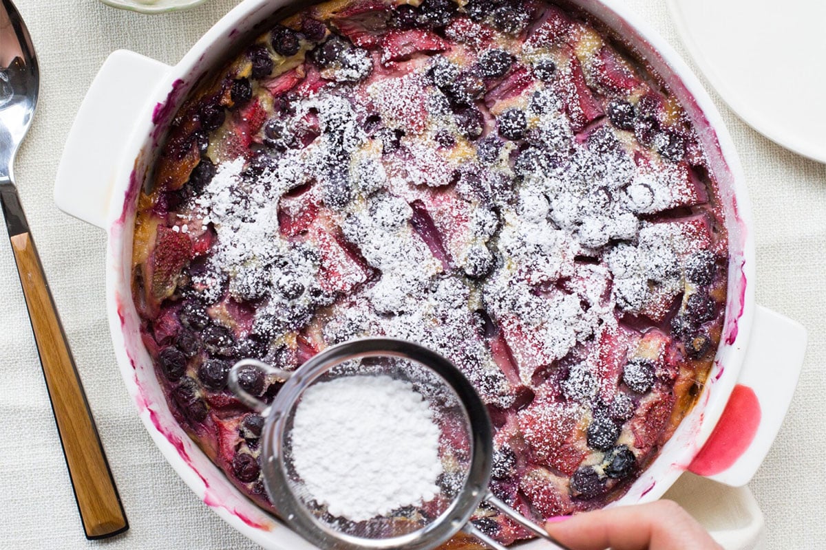Woman sprinkling powdered sugar onto a dish of Summer Berry Clafoutis.