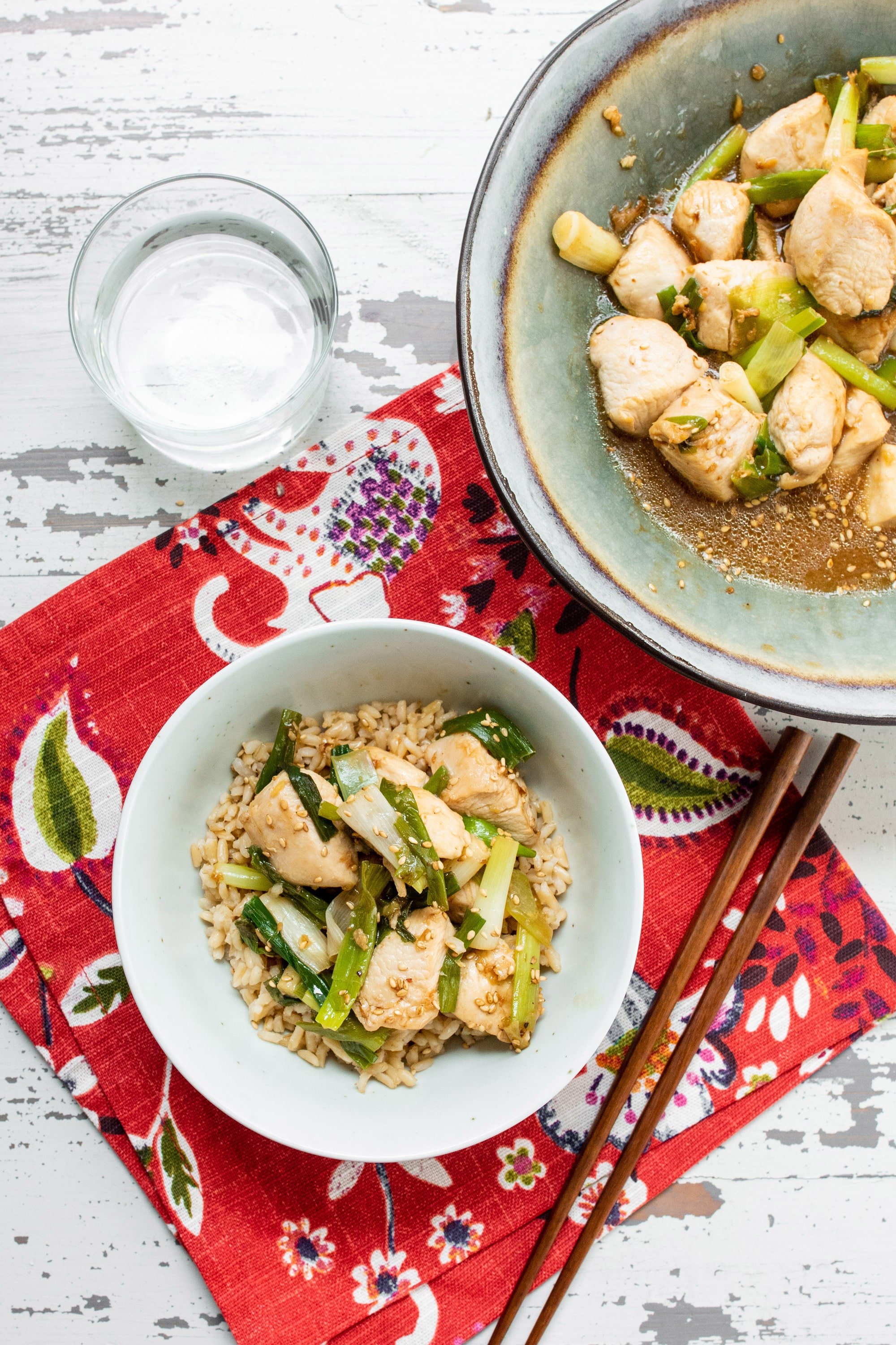 Two bowls of scallion-chicken stir fry on red placemat and white wood table.