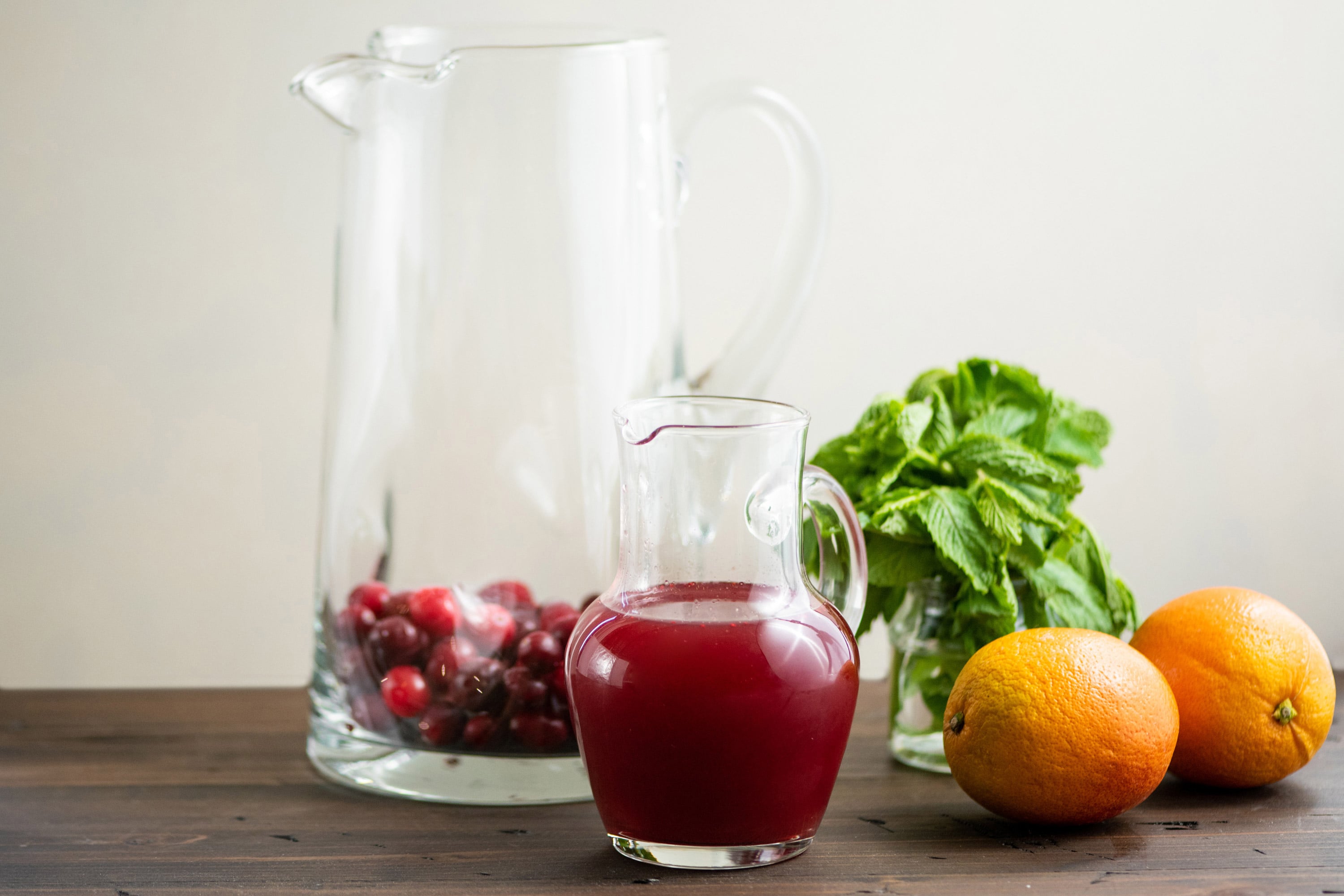 Cranberry Orange Shrub in a small pitcher with a large pitcher with cranberries and some oranges and mint nearby.