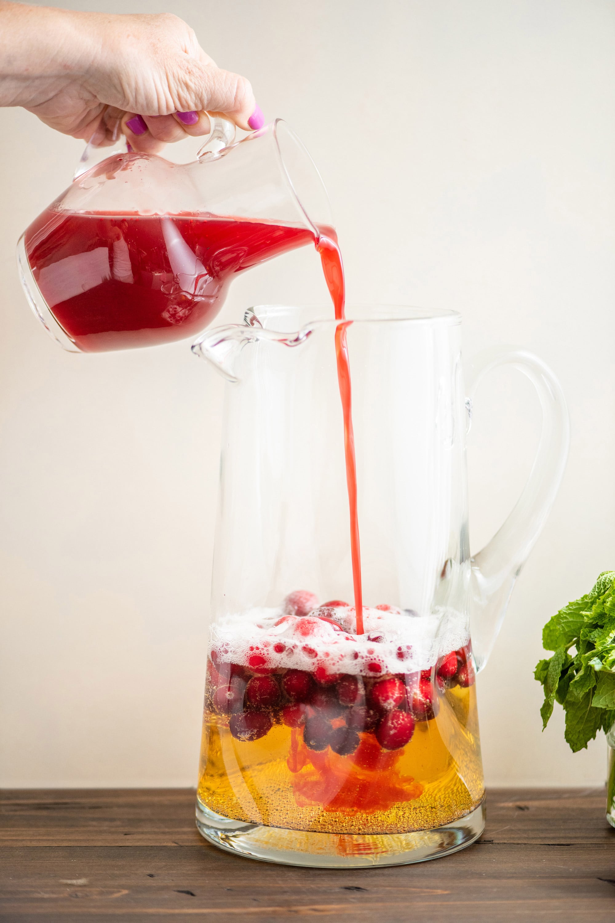 Cranberry shrub being poured into a pitcher with sparkling wine and cranberries in it.