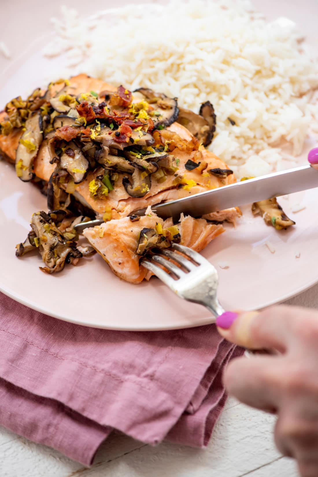 Woman using a fork and knife to cut off a piece of Orange Salmon with Leeks and Mushrooms.