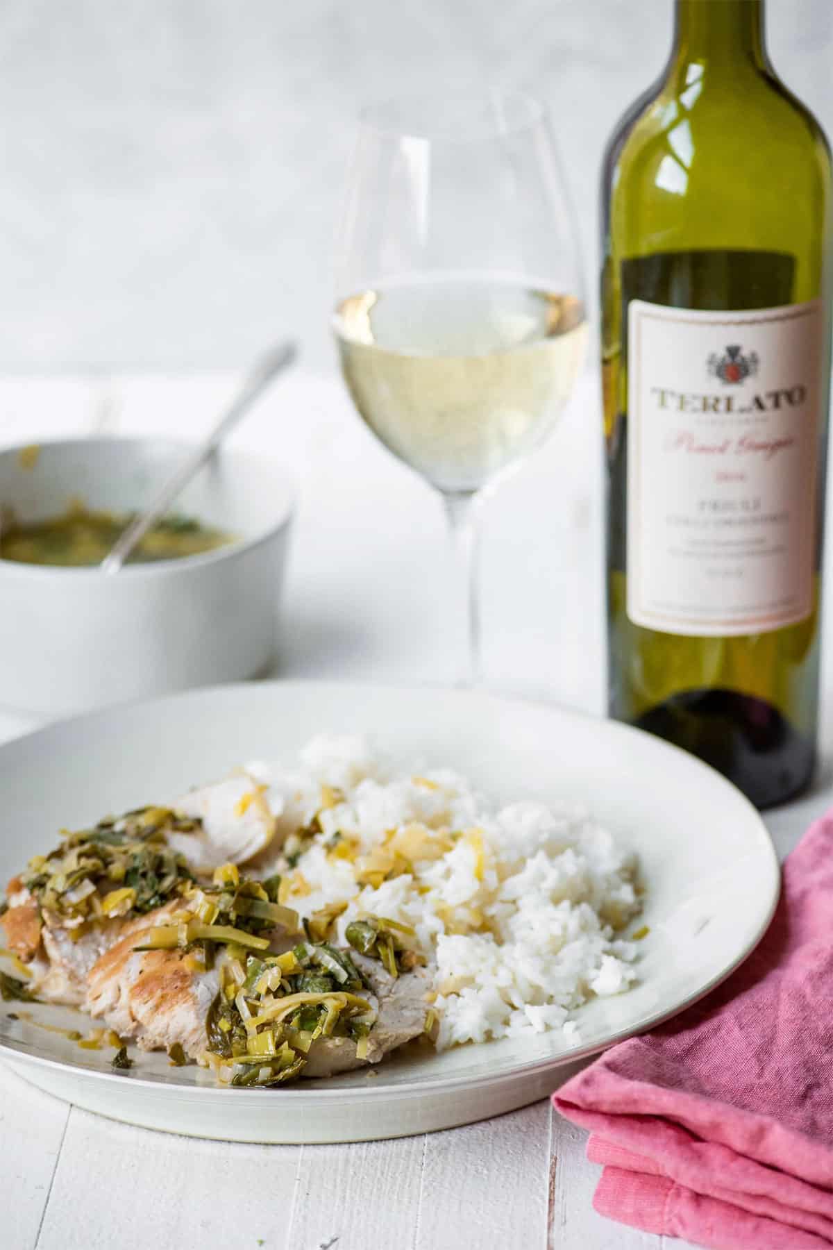 Plate with chicken in white wine and vegetable pan sauce next to wine bottle and glass.