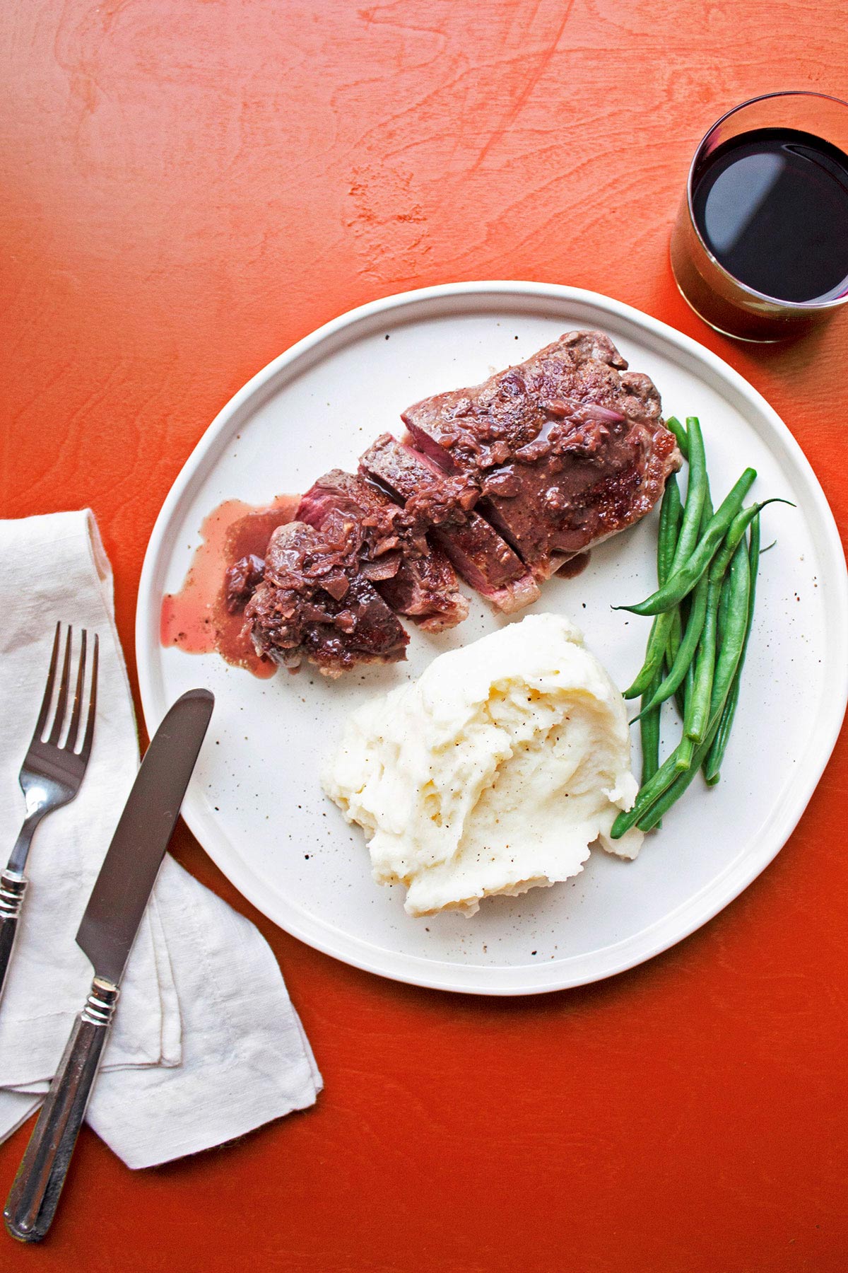 Steak Diane in sauce on white plate with green beans and mashed potatoes on red table.