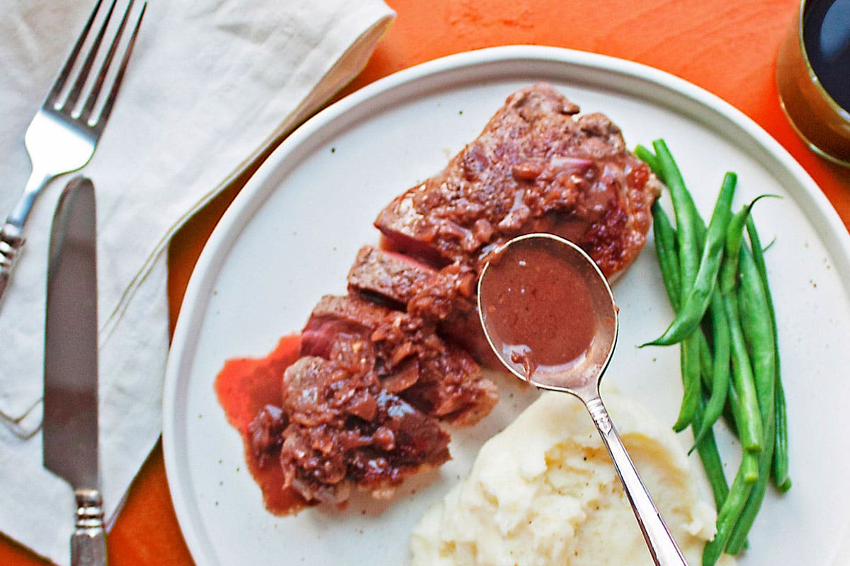 Pouring sauce over strip steak with spoon on plate with sides.