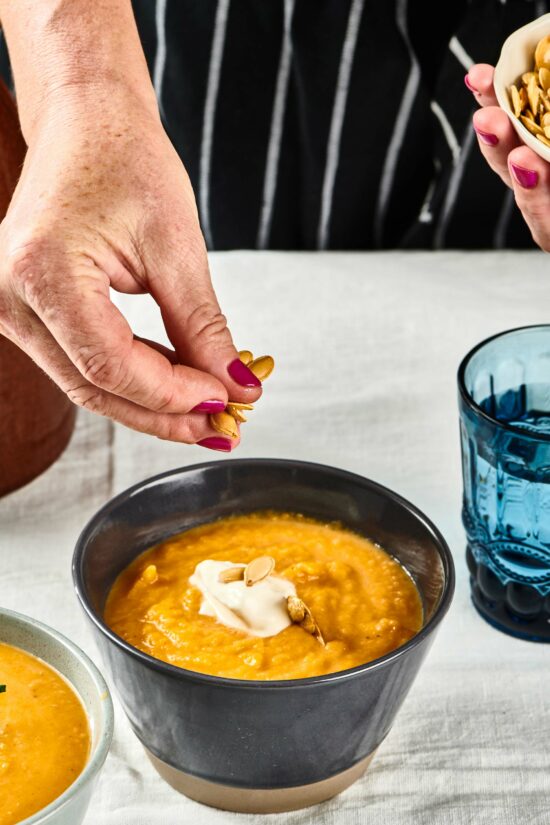 Woman dropping pumpkin seeds into a bowl of Roasted Butternut Squash Soup.