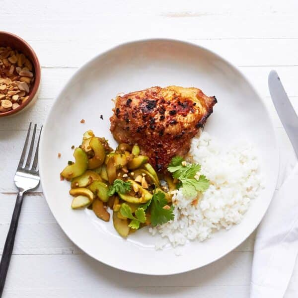 Thai-flavored chicken thigh on white plate with cucumber salad next to bowl of peanuts.