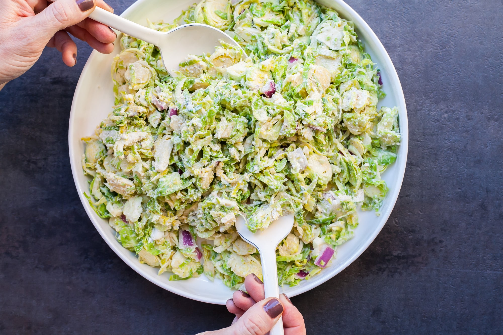 Woman toss creamy Brussels sprouts slaw in a white bowl.