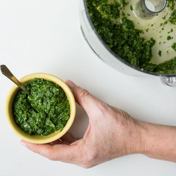 Woman holding yellow bowl of pesto next to food processor.