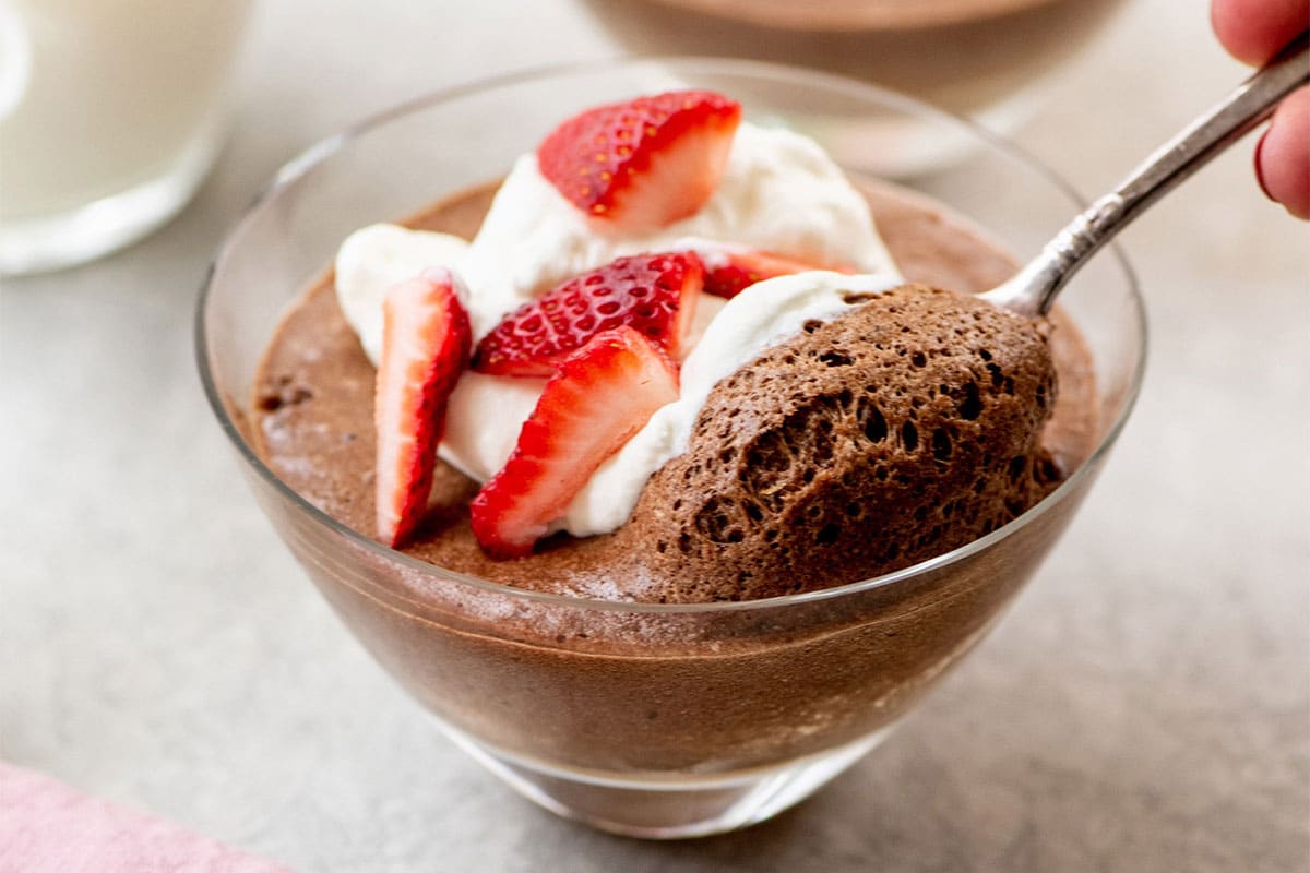 Dipping spoon into homemade chocolate mousse with strawberries and whipped cream out in glass serving dish.