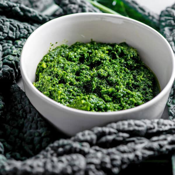 White bowl of Kale Pesto surrounded by fresh kale leaves.