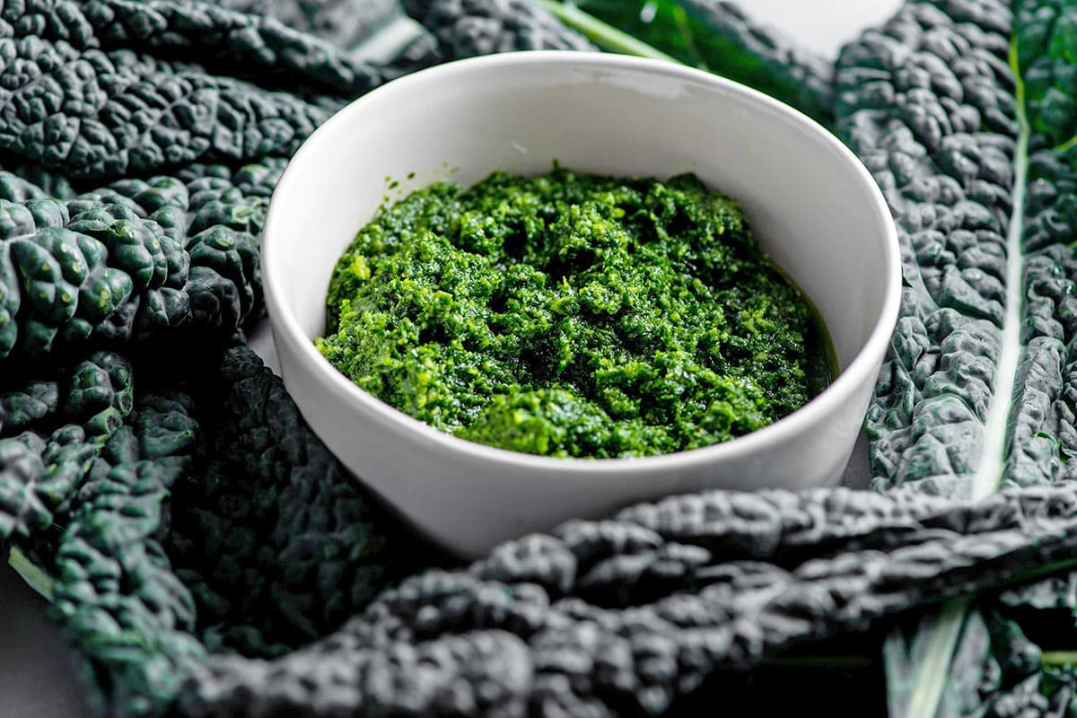 White bowl of Kale Pesto surrounded by fresh kale leaves.