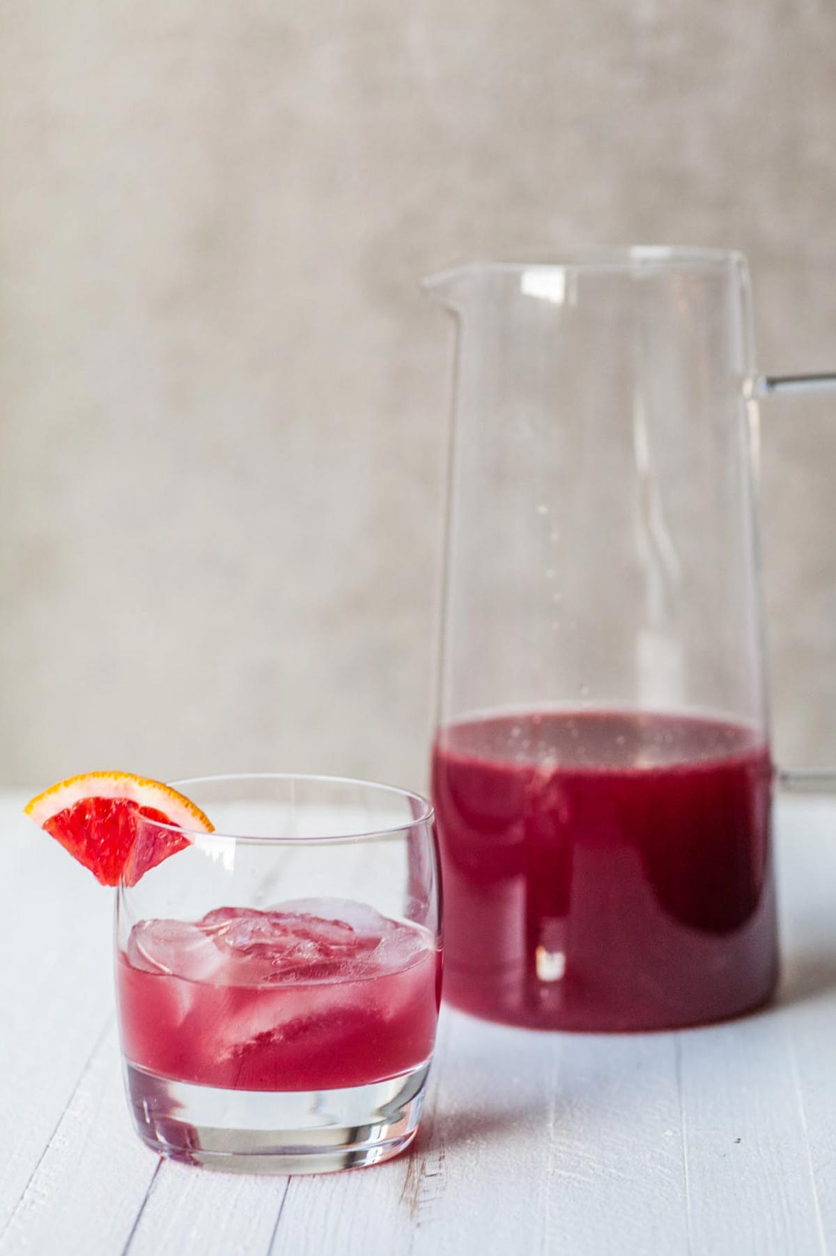 Red pomegranate cocktail in pitcher and ice-filled glass on table.
