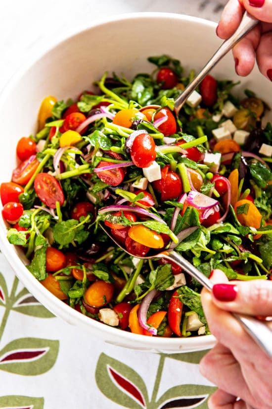 Spoons scooping Tomato, Red Onion, Watercress, Feta and Olive Salad from a bowl.