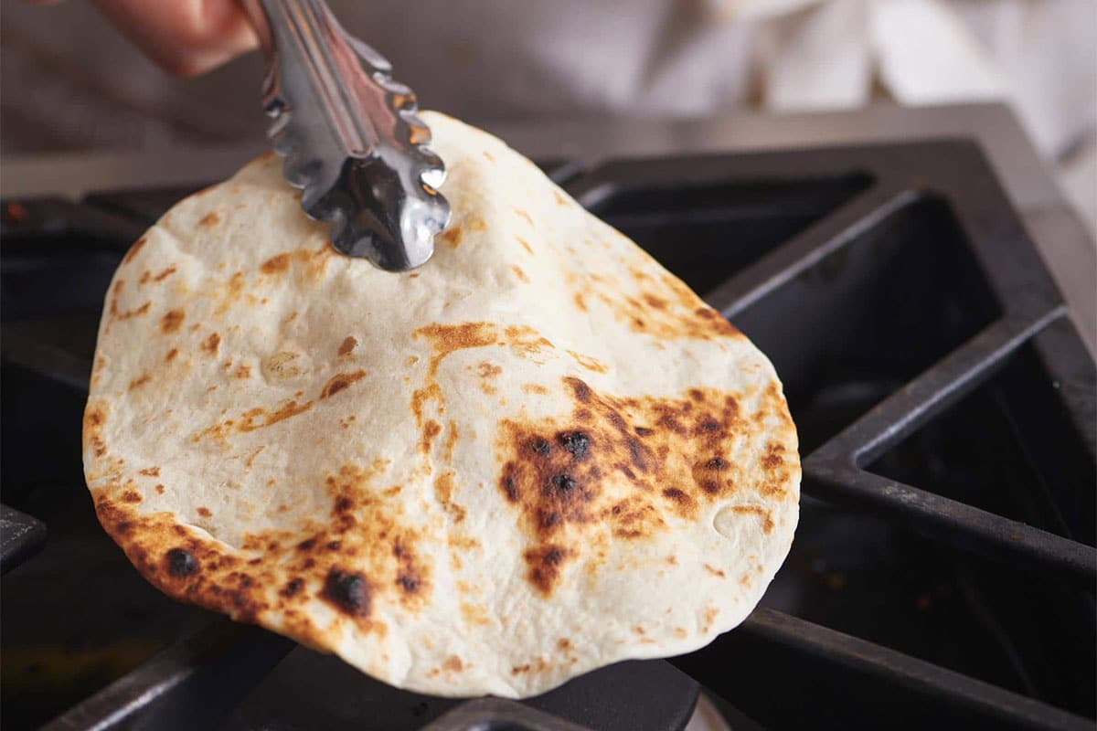 Woman using tongs to flip a tortilla directly on a gas burner.