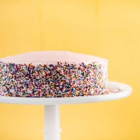 Vanilla cake with pink frosting and sprinkles on pedestal.