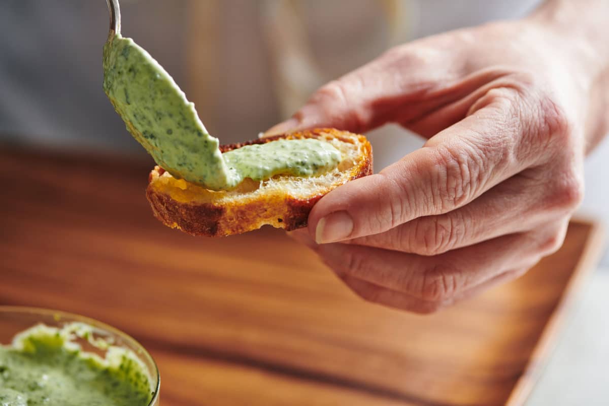 Spoon spreading green Herbed Mayonnaise onto toasted bread.