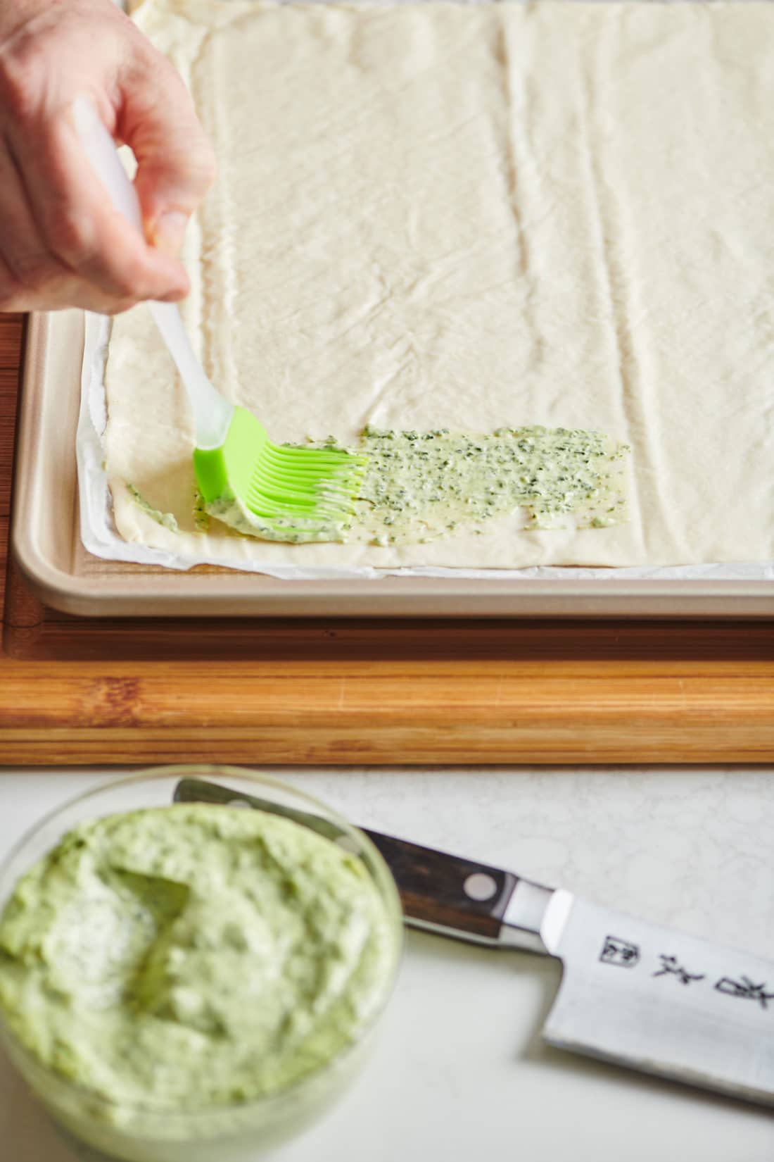 Green brush brushing herbed mayonnaise onto flatbread dough.