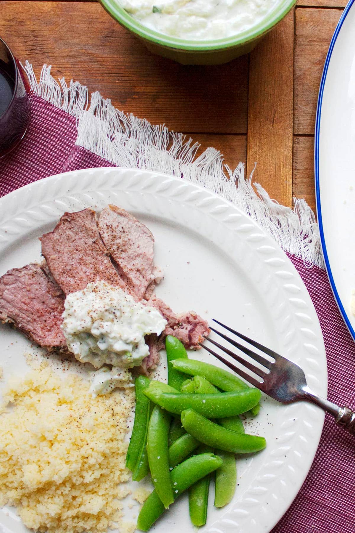 Slices of Mediterranean Leg of Lamb with Tzatziki on a plate with couscous and peas.