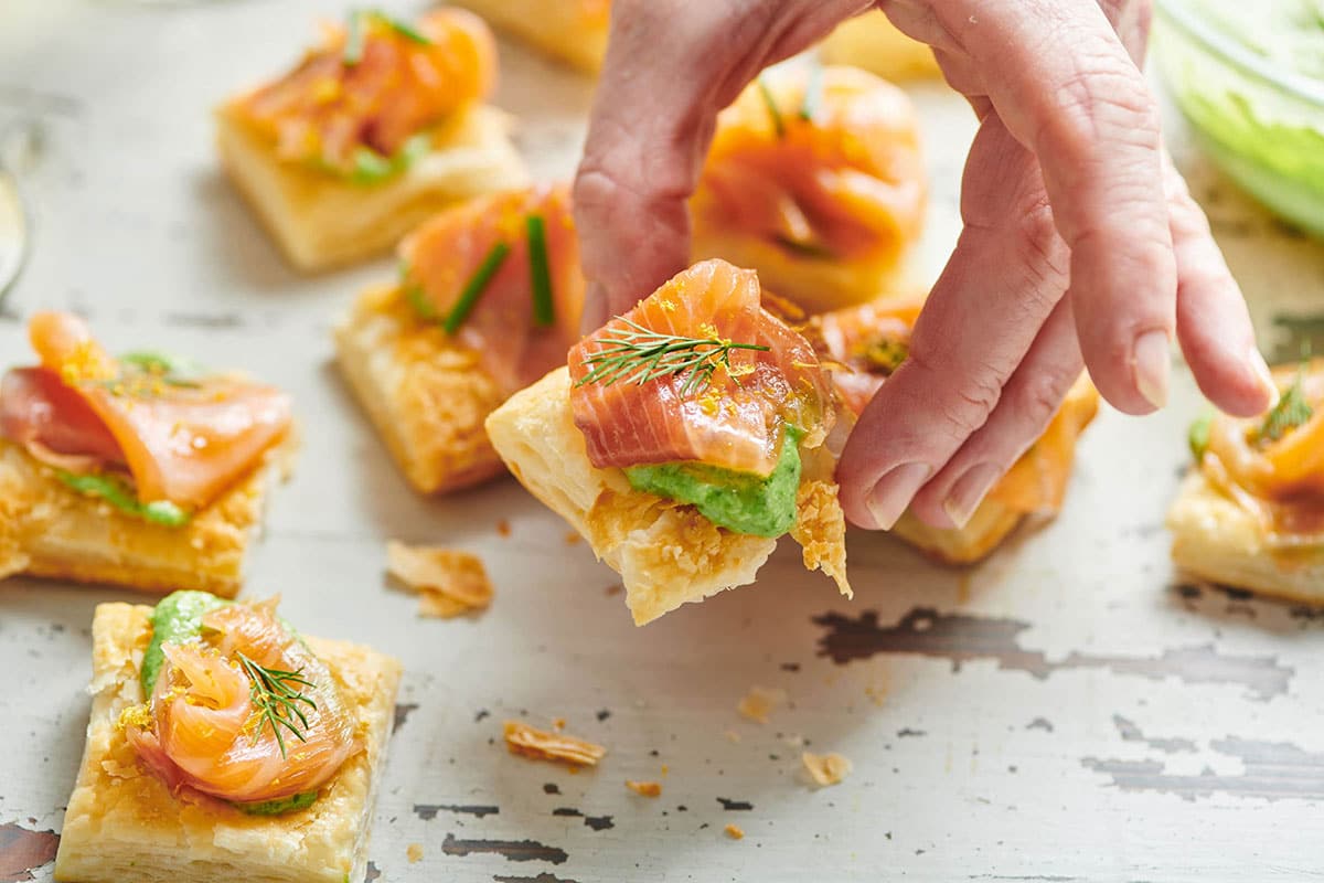 Woman holding small piece of smoked salmon tartlet over table.