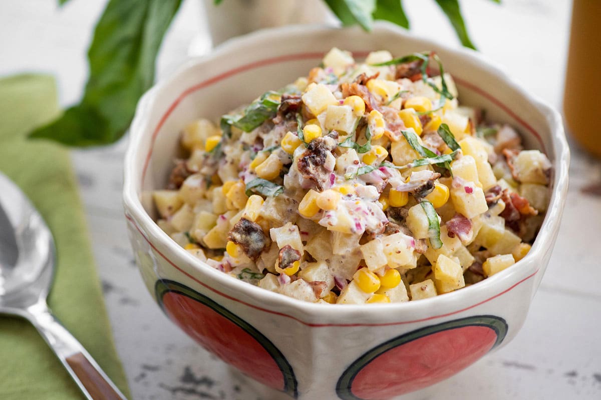 Potato salad with corn and bacon in red and white bowl on table.
