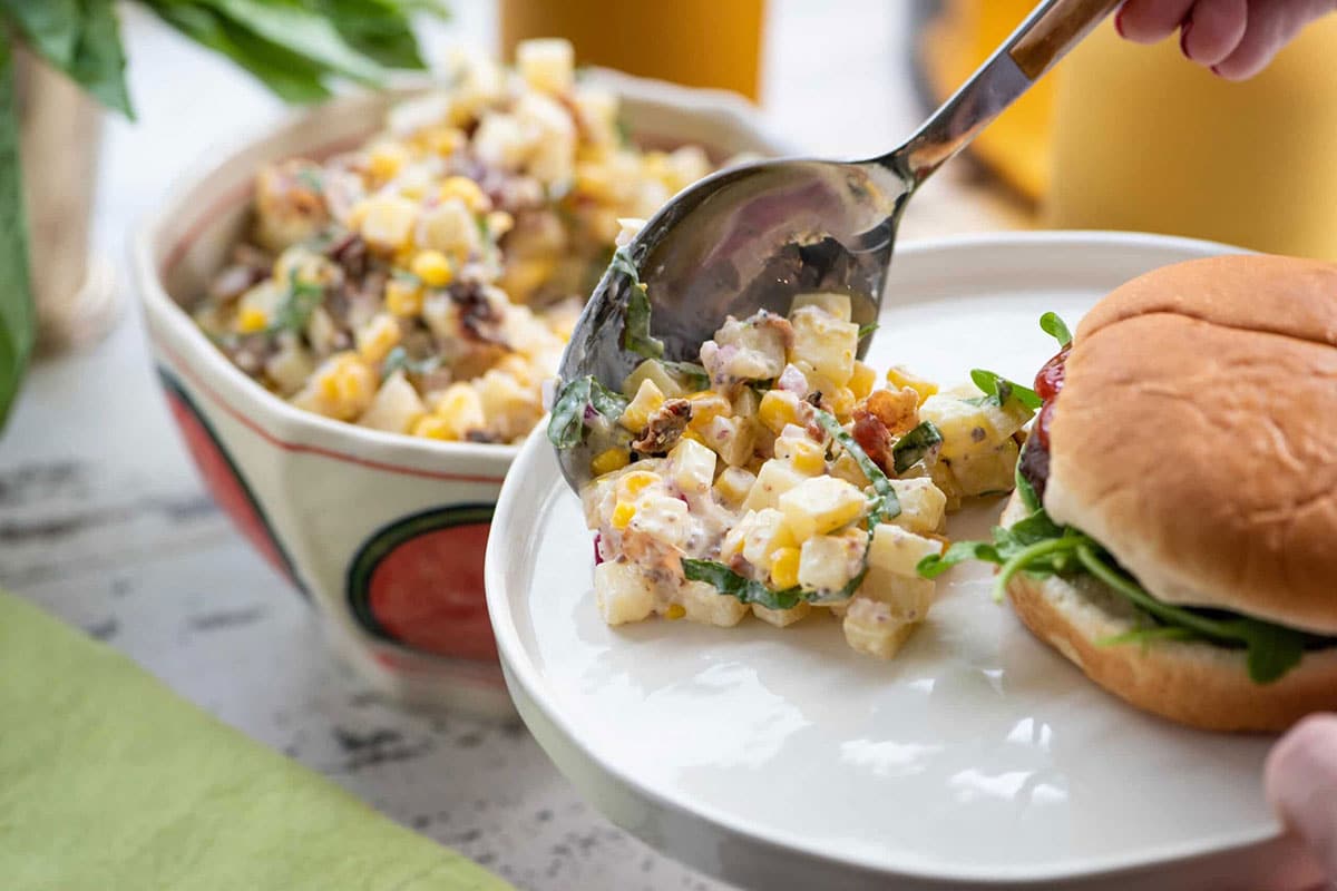 Serving corn potato salad with bacon on plate next to burger.