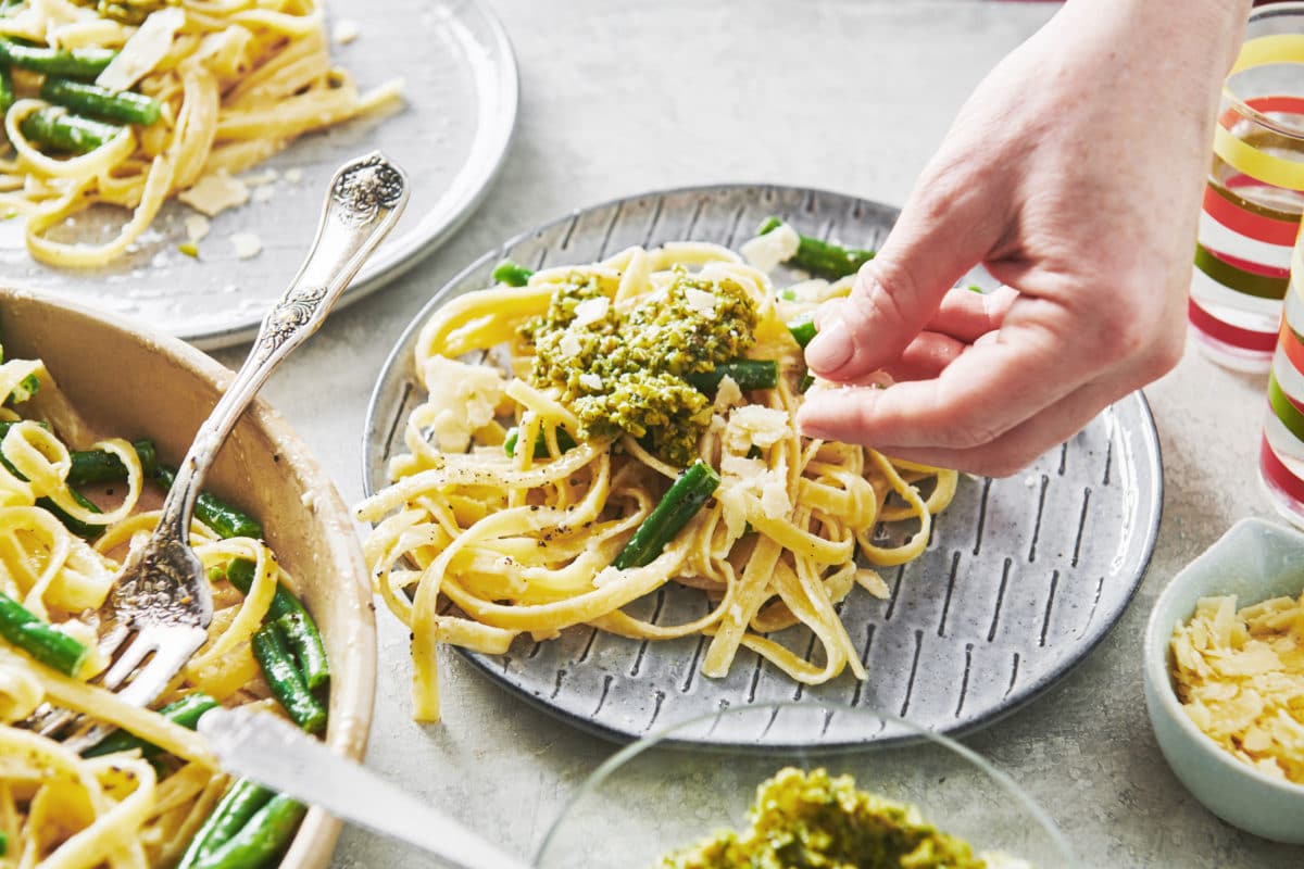 Woman putting parmesan cheese on a plate of Spring Fettuccine Alfredo with Green Beans and Tapenade.