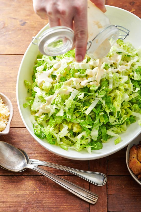 Woman drizzling Caesar dressing over a bowl of romaine lettuce.