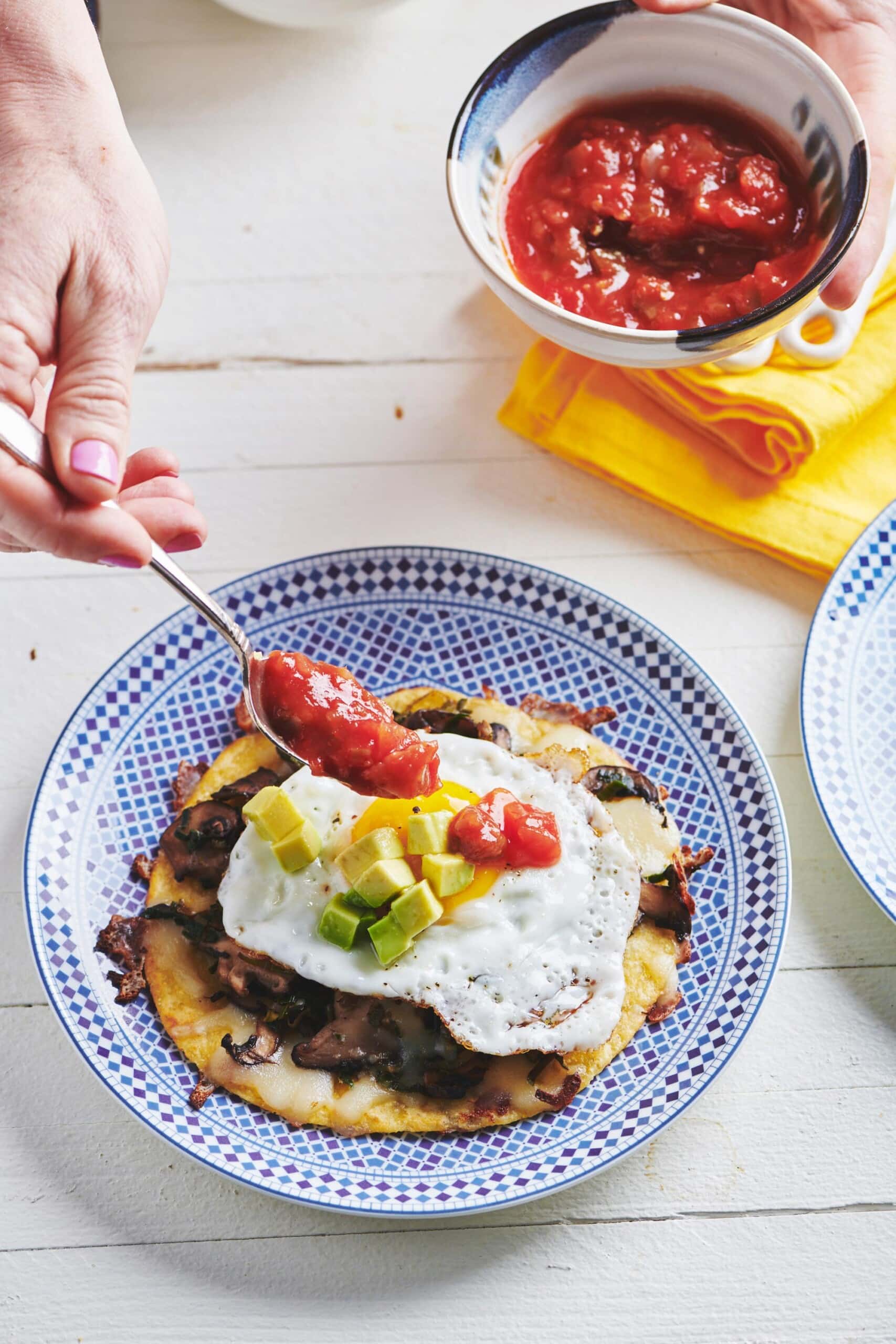 Woman using a spoon to add salsa to a Fried Egg, Mushroom, Leek, and Cheese Tostada.
