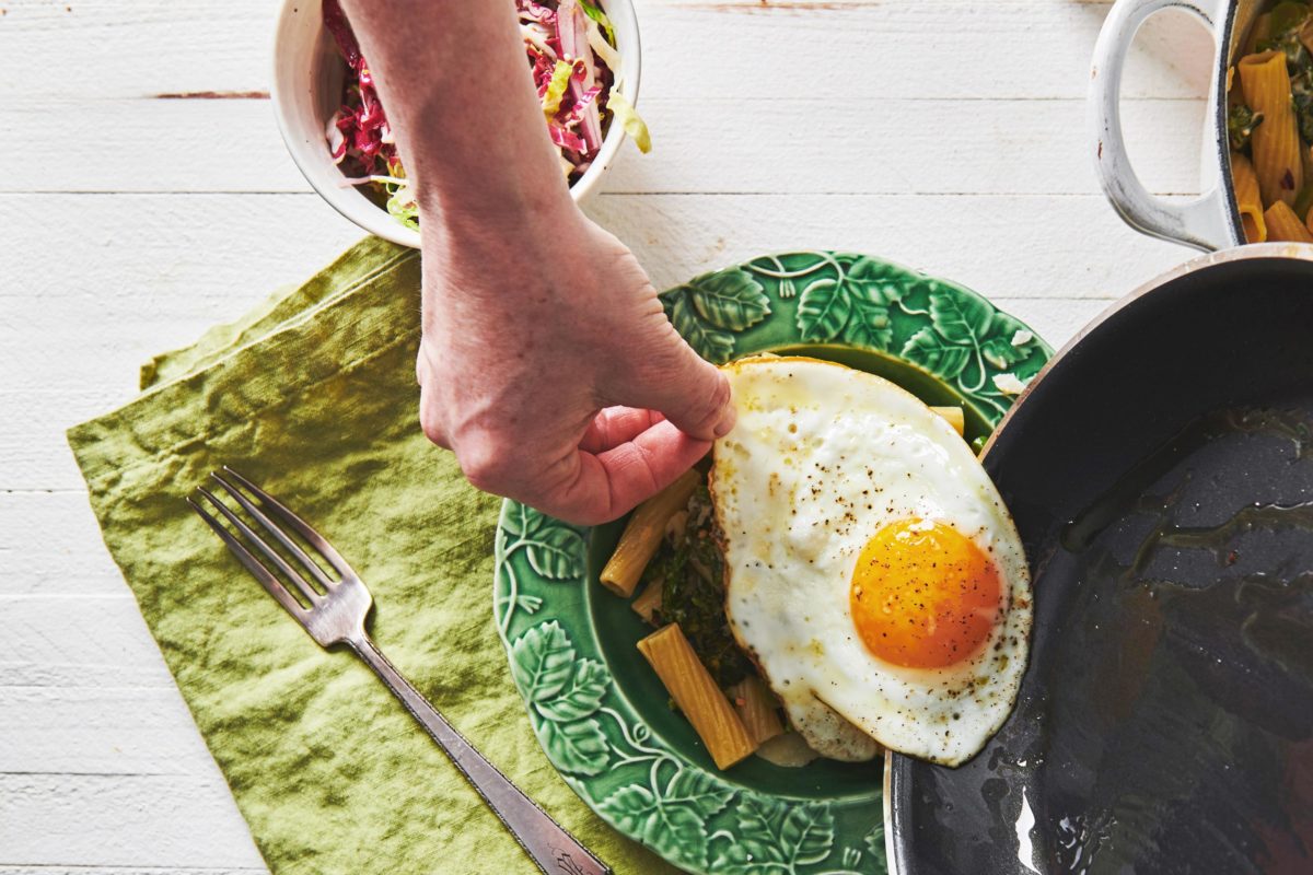 Woman placing a fried egg on top of a plate of Pasta with Feta and Broccolini.