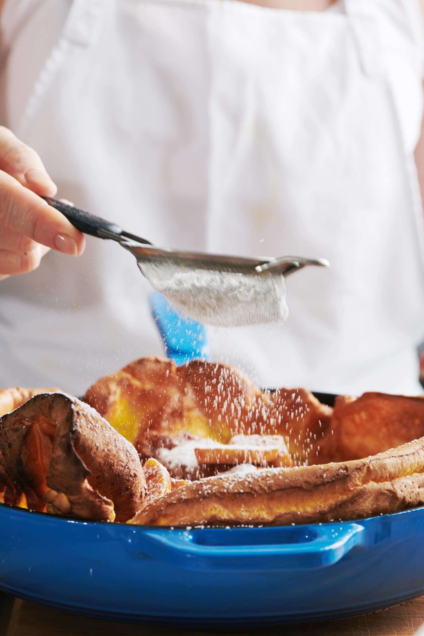 Woman sprinkling powdered sugar onto a Dutch Baby Pancake with Sauteed Apples.