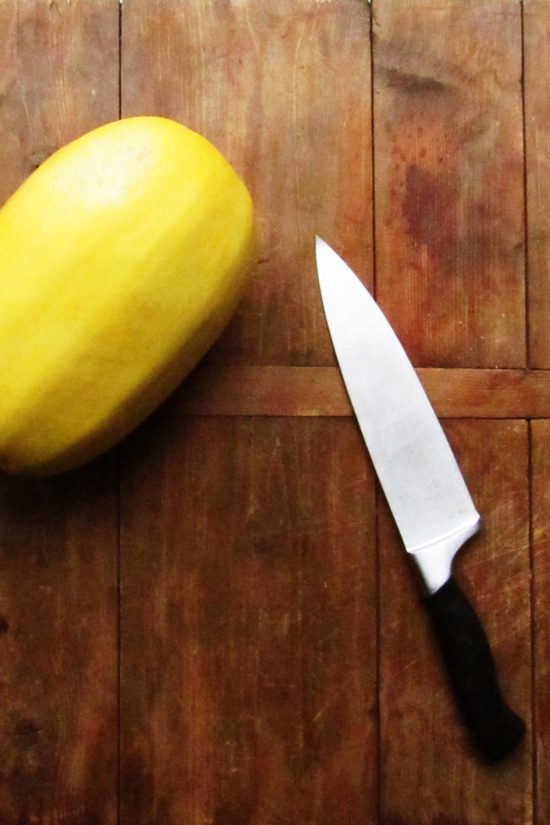 Whole spaghetti squash and a knife on a wooden board.
