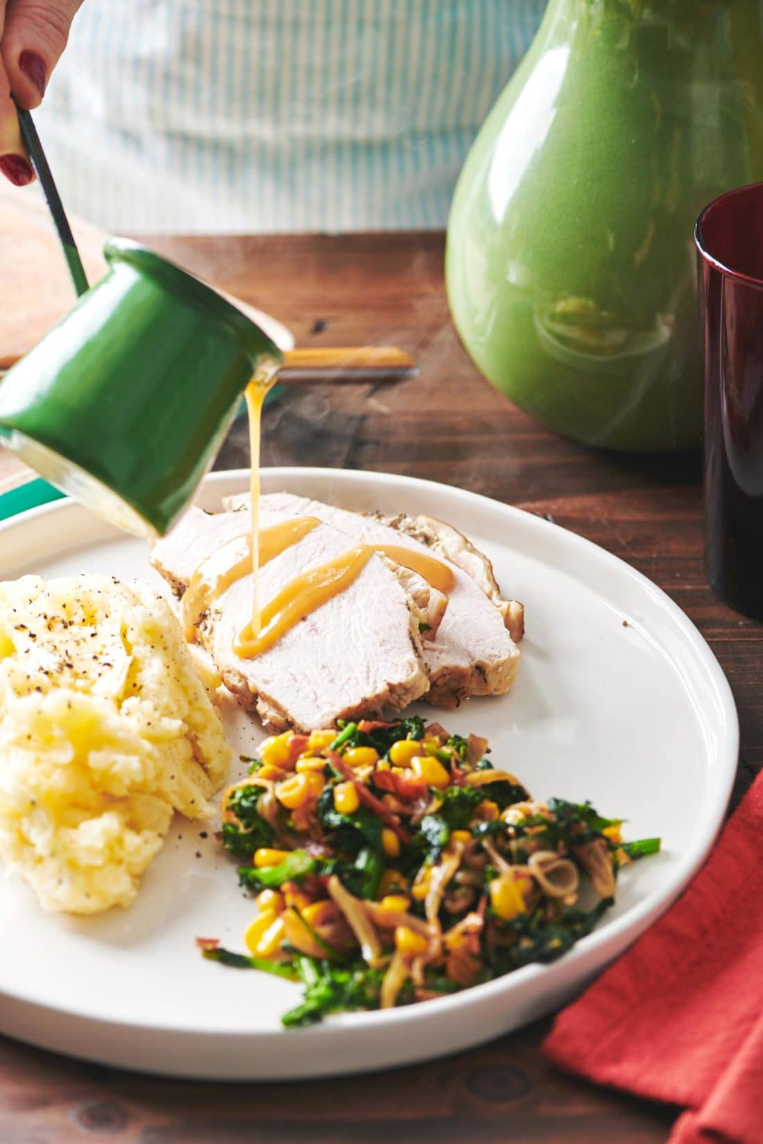 Green cup pouring gravy onto slices of Turkey Breast plated with mashed potatoes and vegetables.